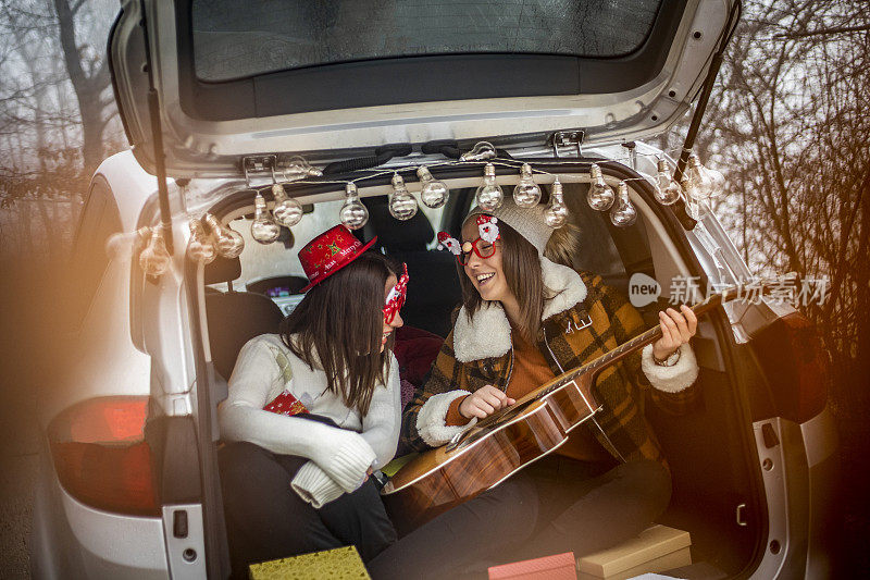 Two women in nature celebrate the new year - Two women in nature celebrate the new year - Two young women celebrate the new year by giving gifts, playing guitar and laugh out - Happy people .两个年轻女性在她们的车里庆祝新年，她们送礼物，弹吉他，放声大笑